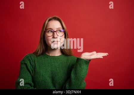 Fröhliche junge Frau in grünem Pullover, die mit der Hand auf leeren Kopierraum zeigt, roter Hintergrund, Mockup. Stockfoto