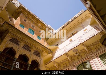 Jaipur, Indien - 15. März 2022: Blick auf die oberen Stockwerke eines Innenhof in einem traditionellen alten Haus mit kunstvoll bemalten Designs, Bogenfenstern mit blauen Fensterläden und architektonischen Details unter klarem Himmel. Stockfoto