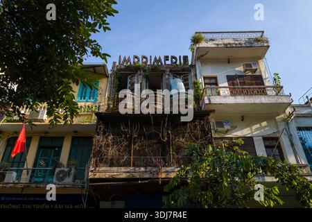 Hanoi, Vietnam - 29. August 2022: Das Bild zeigt alte mehrstöckige Wohngebäude mit Balkonen, ein verblasstes „IMPRIMERIE“-Schild auf dem zentralen Gebäude, sichtbare Klimaanlagen, Pflanzen und eine rote Flagge unter Tageslicht. Stockfoto