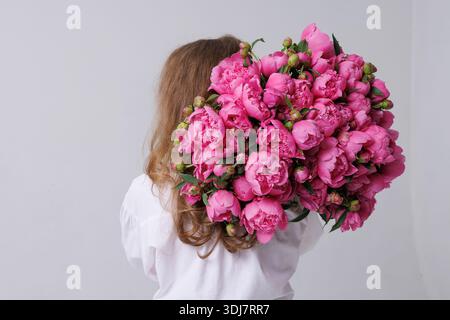 Eine junge Frau mit langen welligen Haaren, die einen großen Strauß rosa Pfingstrosen über der Schulter vor einem neutralen Hintergrund hält. Stockfoto