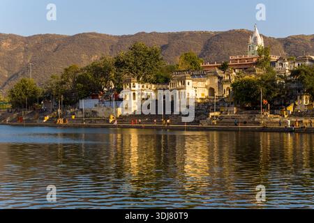 Pushkar, Indien - 17. März 2022: Der Pushkar-See ist mit mehreren Ghats, historischen Gebäuden, Bäumen und einer weißen Kuppelstruktur mit Hügeln im Hintergrund bei klarem Tageslicht sichtbar. Stockfoto