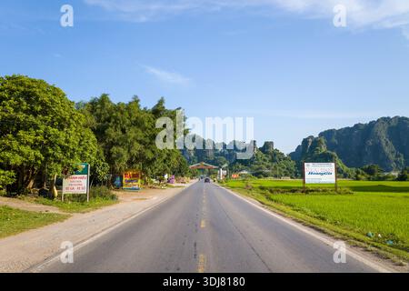 Route de Hanoi vers Mai Chau, Vietnam - 30. August 2022: Das Bild zeigt eine gerade Landstraße, flankiert von grünen Feldern, Straßenbäumen und mehreren Geschäftsschildern, mit Kalksteinbergen im Hintergrund unter einem klaren blauen Himmel. Stockfoto
