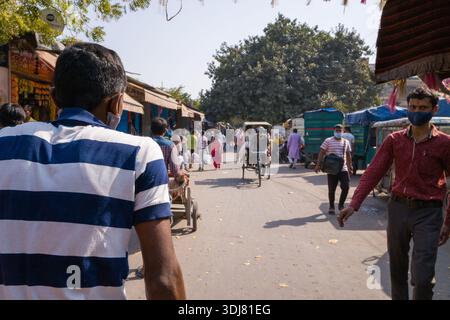 Delhi, Indien - 13. März 2022: Blick von hinten auf einen Rikscha-Fahrer in einem blau-weiß gestreiften Hemd auf einer belebten Straße mit Fußgängern, anderen Rikschas, geparkten Fahrzeugen und Marktständen in Alt-Delhi. Stockfoto