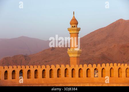 Blick auf die Mauern der Festung Nizwa und die Moschee Al Qalaa vor den Bergen, die von der Nachmittagssonne in Nizwa, Oman, beleuchtet werden. Historische Architektur, warmes Licht, an Stockfoto