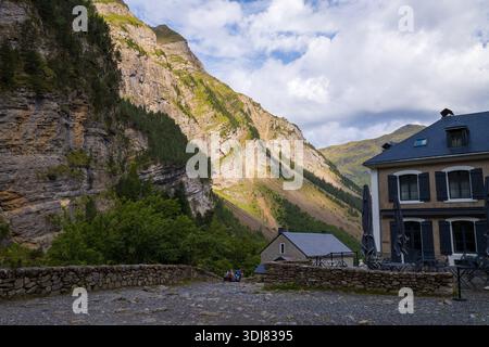 Refuge du Cirque de Gavarnie, Frankreich - 9. August 2022: Ein Blick auf die Berghütten mit einer Steinmauer im Vordergrund, die vor steilen Felswänden und bewaldeten Hängen unter teilweise bewölktem Himmel steht. Stockfoto