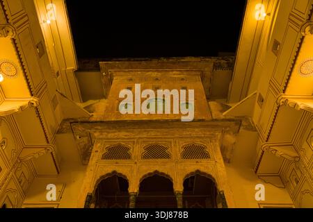 Jaipur, Indien - 16. März 2022: Ein Blick nach oben zeigt die beleuchtete Fassade eines historischen Havelis bei Nacht mit Bögen, dekorativen Gitterleisten, grünen Fensterläden und traditionellen architektonischen Details. Stockfoto