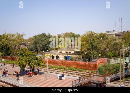 Delhi, Indien - 13. März 2022: Die Menschen sitzen auf Bänken und laufen entlang eines Bahnsteigs neben Bahngleisen, mit einem alten Gebäude, einer roten Backsteinmauer und Bäumen, die im Hintergrund unter klarem Himmel zu sehen sind. Stockfoto