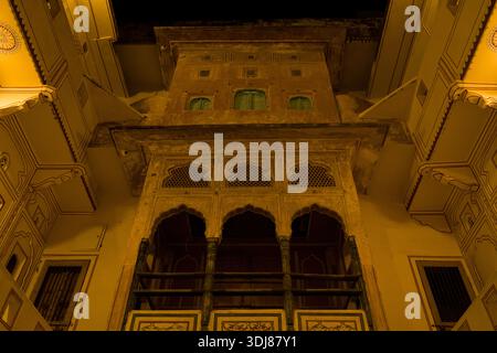 Jaipur, Indien - 16. März 2022: Das Bild zeigt einen Blick nach oben auf eine historische Haveli-Fassade bei Nacht in Jaipur, mit beleuchteten Bögen, dekorativen Gittern und traditionellen architektonischen Details. Stockfoto