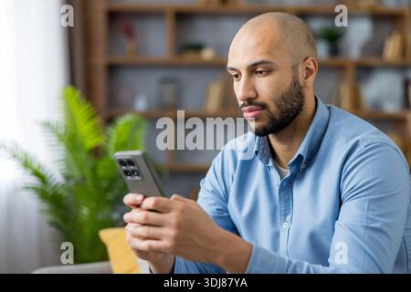 Junger Glatzkopf konzentrierte sich auf den Smartphone-Bildschirm, surfte in sozialen Medien und Messaging-Apps, während er sich in einem modernen Wohnzimmer entspannte, lässige Innenporträts zu Hause Stockfoto