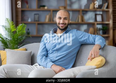 Glücklicher kahlköpfiger Mann mit Bart, blauem Hemd und weißer Hose, der sich auf einer Couch entspannt, auf die Kamera blickt und lächelt, was Komfort und häusliches Leben umfasst Stockfoto