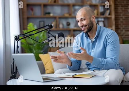 Junger Mann sendet live von zu Hause aus, spricht in ein Mikrofon, während er auf sein Notebook blickt, erstellt Online-Inhalte wie Podcasts, Vlogs oder Streams in einer gemütlichen Umgebung Stockfoto