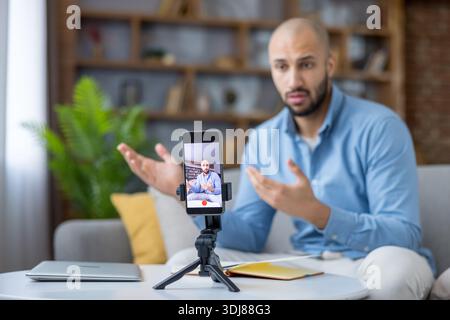 Junger Mann, der zu Hause Online-Inhalte erstellt, sich mit einem Smartphone auf einem Stativ filmt, Informationen weitergibt und mit seinem Publikum über soziale Medien interagiert Stockfoto