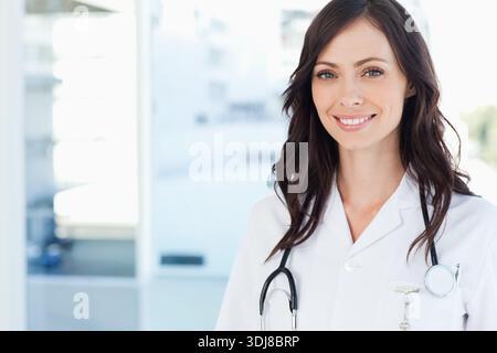 Ärztin stehend, lächelnd in der Klinik mit Laborkittel, Stethoskop und Ausweis, Kopierraum. Arzt, Gesundheitswesen, Büro, sonnenbeleuchtet, städtisch, Hochformat, PR Stockfoto