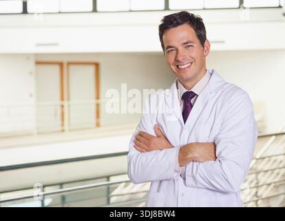 Männlicher Arzt steht mit überkreuzten Armen im Krankenhausatrium, trägt weißen Labormantel, Kopierraum. Arzt, Kopfschuss, professionell, medizinisch, Klinik, Bogen Stockfoto