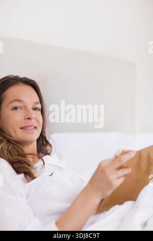 Frau, die sich auf dem Bett liegend, mit weißem Pyjamaoberteil, braunem Buch und Blick auf die Kamera. Schlafzimmer, Kopfteil, Bettwäsche, Notizbuch, Lesen, ruhig, c Stockfoto
