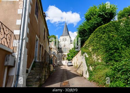 Pouilly sur Loire, Frankreich - 27. Juni 2022: Eine schmale Straße, die von Wohnhäusern und einer grünen Steinmauer gesäumt ist, führt zu einer historischen Steinkirche mit einem hohen Schieferturm unter blauem Himmel mit verstreuten Wolken. Stockfoto
