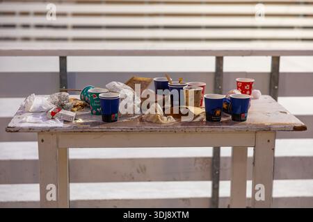 VINKOVCI, KROATIEN, 12.27.2025: Ein verwitterter Holztisch im Freien mit Einwegbechern, zerknitterten Papieren, Verpackungen, Servietten und Essensresten. T Stockfoto