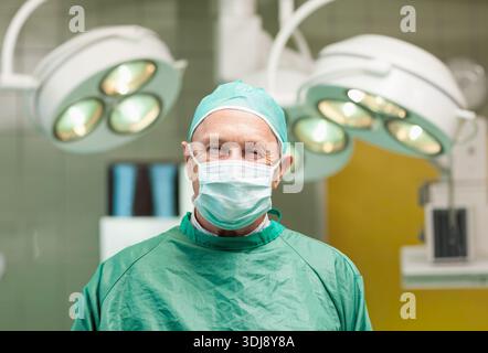 Senior männlicher Chirurg stehend, mit grünem OP-Kleid und Maske im Operationssaal unter Licht. Arzt, Scrubs, steril, Krankenhaus, medizinisch, clin Stockfoto