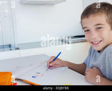 Junge, der in der Küche sitzt, zeichnet in Notizbuch mit blauem Stift und orangefarbenem Bleistiftetui, Kopierraum. Kind, Zeichnen, Skizzenbuch, Schreibwaren, bunt, modern, l Stockfoto