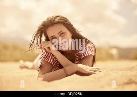 Nahaufnahme einer lächelnden Frau, die entspannt auf dem Sand liegt und die sonnige Strandatmosphäre genießt, unbeschwerte Momente, Sonnenschein, exhuberantes Lächeln genießt, Stockfoto