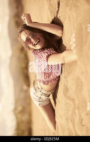 Frau liegend auf dem Sand, lächelnd, in lässigem gestreiftem Hemd und Sonnenbrille am Strand, fröhliches Glück, überglücklich sein, Entspannung, Wellness, holi Stockfoto