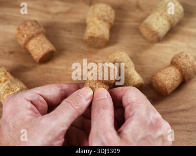 Ein Mann, der einen einzigen benutzten Sektkorken in den Händen hält, und mehrere weitere auf einem Holztisch im Hintergrund verstreut. Stockfoto