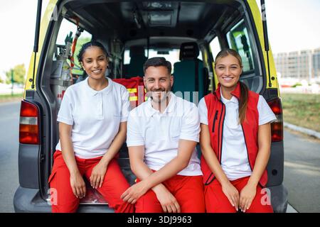 Sanitäter und Arzt stehen hinter dem Krankenwagen. Der Arzt trägt einen medizinischen Traumabeutel. Gruppe von drei Sanitätern, die vor dem Krankenwagen stehen Stockfoto