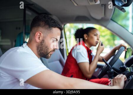 Zwei Sanitäter vor einem Krankenwagen. Der Fokus liegt auf dem Mann, einem jungen Mann im Alter von 20 Jahren, der durch das offene Fenster auf der Fahrerseite schaut. Stockfoto