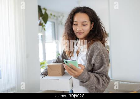 Lächelnde junge asiatische Frau beim Online-Shopping mit Smartphone zur Hand, die ein zugestelltes Paket per Zustellungsdienst erhält. Online-Shopping, Online-Bank Stockfoto