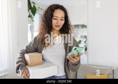 Lächelnde junge asiatische Frau beim Online-Shopping mit Smartphone zur Hand, die ein zugestelltes Paket per Zustellungsdienst erhält. Online-Shopping, Online-Bank Stockfoto