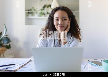 Youn asiatischer Arzt, der Laptop benutzt und am Schreibtisch sitzt. Ärztin mit weißem Mantel und Stethoskop, die am Computer am Arbeitsplatz arbeitet Stockfoto