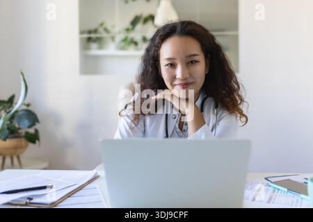 Youn asiatischer Arzt, der Laptop benutzt und am Schreibtisch sitzt. Ärztin mit weißem Mantel und Stethoskop, die am Computer am Arbeitsplatz arbeitet Stockfoto