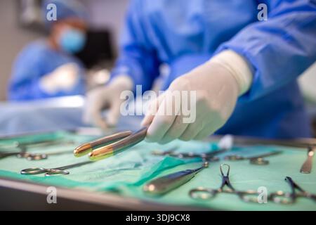 Nahaufnahme einer Hand mit Handschuhen, die ein chirurgisches Instrument aus einem sterilen Tablett aufnimmt. Verschiedene chirurgische Werkzeuge auf einem grünen Tuch im OP Stockfoto