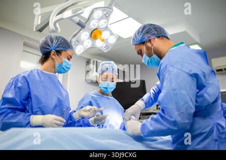 Chirurgen und Helfer arbeiten während der Operation zusammen. Ärztliches Team in sterilen Peelings, das einen Eingriff unter hellem Licht durchführt. Stockfoto