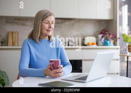 Seniorin lernt und arbeitet von zu Hause aus, sieht Inhalte auf einem Laptop an und genießt eine Tasse Kaffee am Küchentisch. Moderne Technik ist dabei dabei dabei Stockfoto