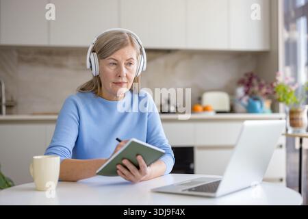 Ältere Frau zu Hause mit Kopfhörern, konzentriert sich auf Laptop, macht Notizen während eines Online-Kurses oder Videoanrufs, die sich mit Fernunterricht, Arbeit oder Selbstverbesserungsaktivitäten beschäftigen Stockfoto