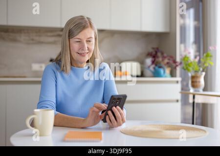 Seniorin sitzt an einem weißen Küchentisch, hält und interagiert mit ihrem Handy und genießt an einem entspannten Morgen zu Hause eine warme Tasse Kaffee Stockfoto