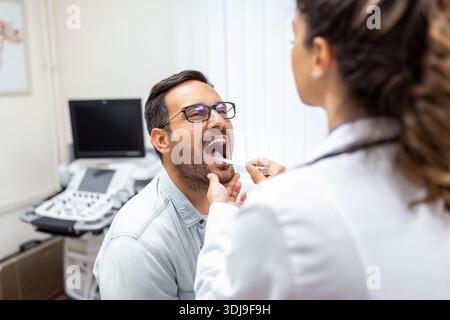 Arzt mit Inspektionsspachtel zur Untersuchung des Rachens des Patienten. HNO-Arzt, der eine Rachenuntersuchung durchführt. Der Patient öffnete seinen Mund zur Kehle Stockfoto