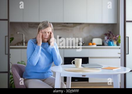 Ältere Frau mit intensiven Kopfschmerzen, am Küchentisch sitzen und ihre Schläfen drücken, Migräne-Symptome bekämpfen und sich zu Hause erschöpft fühlen Stockfoto