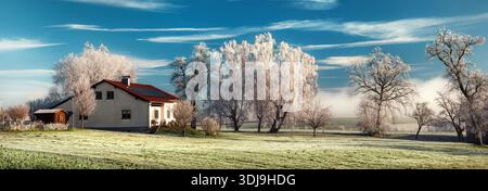 Ländliches Haus in magischer gefrorener Landschaft. Panoramablick auf das Winterwunderland mit Haus, blauem Himmel und Raureif auf Bäumen und Wiese Stockfoto