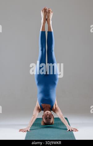 Reife Frau in blauer Activewear, die im Innenbereich Kopfstand-Yoga-Pose auf der Matte macht und Stärke und Balance demonstriert. Stockfoto