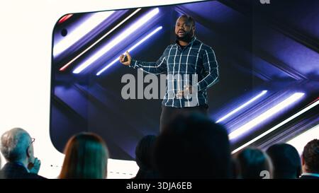 Der Senior Designer von Black stellt den Teilnehmern während der Podiumsdiskussion des Business Summit ein neues Smartphone vor. Produktpräsentation für Elektronik mit Vorstellung neuer Mobilgeräte. Stockfoto