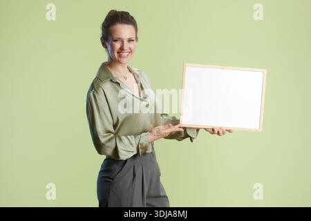 Eine fröhliche Frau mittleren Alters präsentiert ein weißes Zeichen in einer hellgrünen Seidenbluse. Sanfte Beleuchtung verbessert den positiven und professionellen Kontext. Stockfoto
