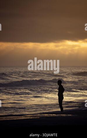 Eine Person hat eine Silhouette am Strand, als Wellen bei Sonnenuntergang am 18. Dezember 2025 auf Dauphin Island, Alabama, abstürzen. (Foto: Carmen K. Sisson/Cloudybright) Stockfoto
