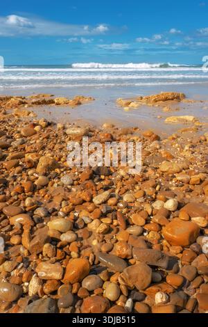 Die felsige Küste übergeht in flaches Wasser und brechende Wellen, unter einem hellblauen Himmel mit verstreuten Wolken Stockfoto