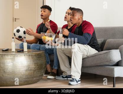 Verschiedene männliche Freunde auf der Couch sehen sich das Spiel an, tragen passende Shirts, die Fußball und Chips halten Stockfoto