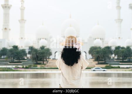 Touristenfrau, die die herrliche Scheich-Zayed-Moschee in Abu Dhabi mit Blick auf den See und üppigen grünen Bäumen im Vordergrund betrachtet Stockfoto