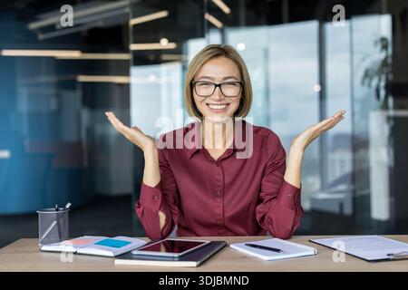 Lächelnde junge Geschäftsfrau mit Brille hebt die Hände in einer feierlichen Geste und drückt Freude und Selbstvertrauen aus, während sie an ihrem Schreibtisch sitzt Stockfoto