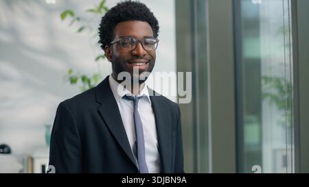 Afroamerikanischer selbstbewusster Geschäftsmann lächelt in die Kamera glücklicher Blick aus dem Fenster erfolgreicher Geschäftsmann, der sich positiv im modernen Büro fühlt männlicher Mann Stockfoto