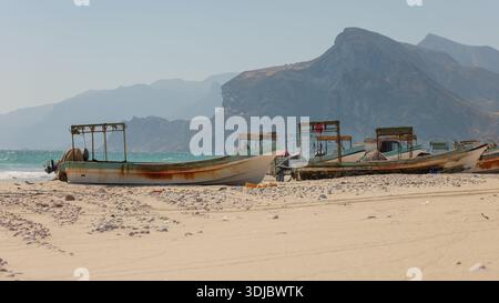 Mehrere verwitterte Fischerboote ruhen an einem Sandstrand in Oman, mit Kieselsteinen und türkisfarbenen Meereswellen in der Nähe und majestätischen, trüben Bergen i Stockfoto